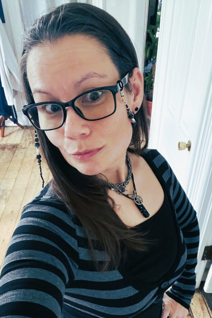 Adult woman with long dark hair wearing black glasses and a striped top, taking a cozy indoor selfie with soft lighting and layered necklaces.